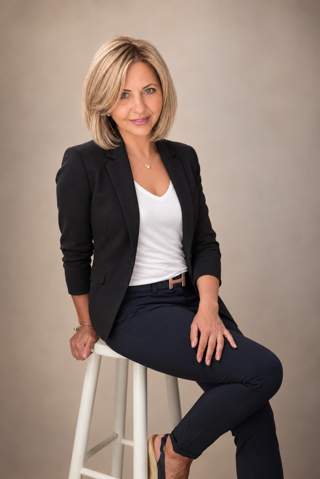 Oksana Huber im schwarzen Blazer auf einem hellen Hocker vor warmem Hintergrund.